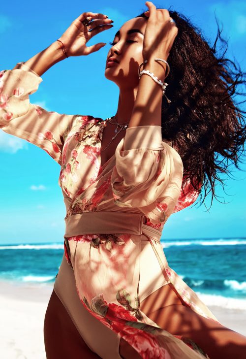 portrait of beautiful caucasian sunbathed woman model with dark long hair in beige flying dress posing on summer beach with white sand on blue sky and ocean background. Touching her hair